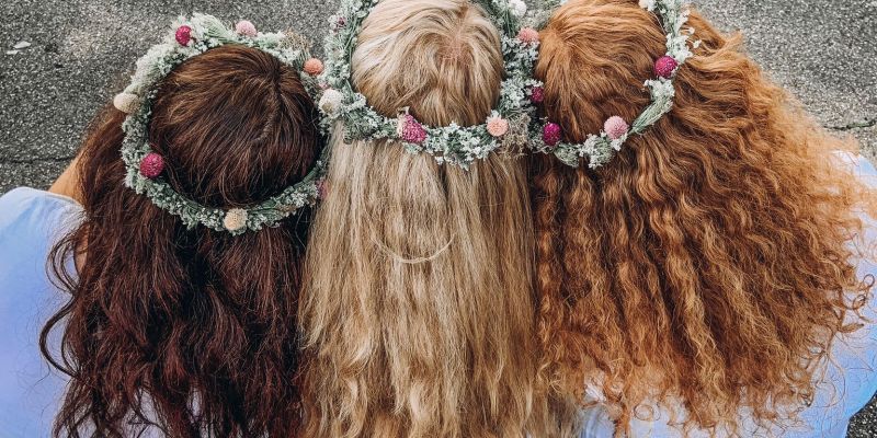 Das Bild zeigt drei Köpfe mit Blumenkränzen in langen, welligen Haaren von hinten.